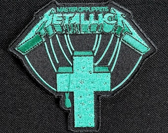 May include: A black patch with the words "MASTER OF PUPPETS" and "METALLICA" in green, with a green cross design. The patch has a black border and is likely an iron-on or sew-on patch for clothing or accessories.