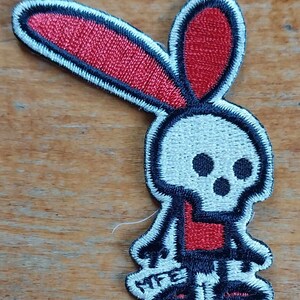Pode incluir: Etiqueta bordada com uma caveira branca com olhos pretos e boca vermelha, vestindo uma camisa vermelha e calças pretas. A caveira tem uma orelha de coelho vermelha no topo.