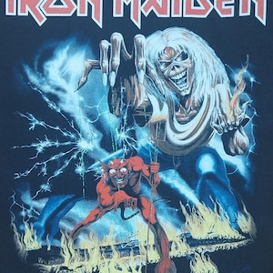 Può includere: T-shirt nera con la scritta rossa "Iron Maiden". La grafica raffigura una figura scheletrica con capelli bianchi e un diavolo rosso davanti alle fiamme, con fulmini sullo sfondo.