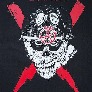 Op de afbeelding: Zwarte afbeelding met een witte schedel met een helm, een rood symbool en gekruiste rode bliksemschichten. De tekst "S.O.D." staat in het rood bovenaan. De schedel heeft zwarte details.