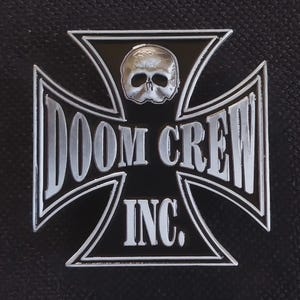 May include: A black and silver metal pin shaped like an iron cross. The design features a silver skull at the top and the text DOOM CREW INC. in silver lettering across the center and bottom sections against a black enamel background.