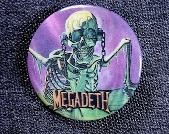 May include: Round button with a purple and green graphic of a skeleton wearing headphones. The skeleton has a skull and crossbones design and the word "MEGADETH" is printed in gold.