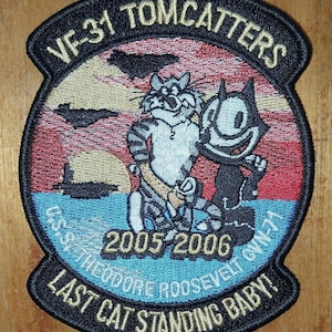 May include: A black and white embroidered patch with the text "VF-31 TOMCATTERS" in a black circle. The patch features a cartoon cat with a striped shirt and a black cat with a white shirt. The text "USS THEODORE ROOSEVELT CVN-71" and "LAST CAT STANDING BABY!" are also on the patch. The years "2005 2006" are in the centre of the patch.