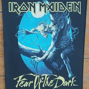 May include: Black rectangular patch featuring the band name "Iron Maiden" in yellow block letters above a blue and black illustration. The illustration depicts a skeletal figure with a menacing expression. The text "Fear Of The Dark" is below the illustration.