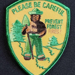 May include: A yellow and green embroidered patch with a cartoon ranger bear holding a shovel and walking stick. The text "PLEASE BE CAREFUL PREVENT FOREST FIRES!" is embroidered on the patch.
