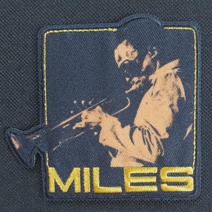 Könnte beinhalten: Dunkelblauer Aufnäher mit goldfarbenem Rand, der einen Musiker in Sepia zeigt, der Trompete spielt. Der Name "MILES" ist in goldenen Buchstaben auf den Aufnäher gestickt.