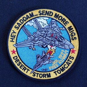 以下が含まれることがあります： 「Hey Saddam Send More Migs Desert Storm Tomcats Jan 1991 Free Kuwait」というテキストと戦闘機が描かれた黄色と青の刺繍パッチ。