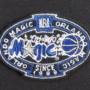 Puede incluir: Un parche bordado azul y blanco con el texto "Orlando Magic" y "NBA" en un círculo. El parche también incluye un balón de baloncesto y el texto "Since 1997".