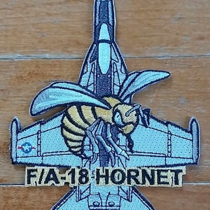 May include: Embroidered patch featuring an F/A-18 Hornet fighter jet design with a stylized bee. The patch is primarily light blue, black, and yellow, with the text "F/A-18 HORNET" in a rectangular banner.