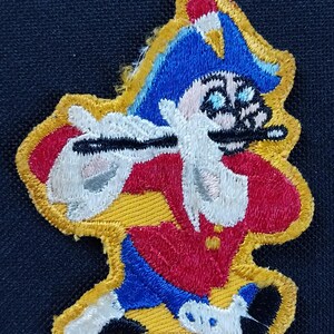 May include: Embroidered patch featuring a cartoon character in a red jacket, blue pants, and a blue hat with a white feather. The character has a white beard and is holding a black object. The patch has a yellow border and is set against a dark blue background.