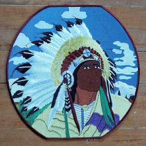 May include: Embroidered patch of a Native American chief wearing a colorful headdress with feathers. The chief is looking to the right and has a serious expression on his face. The background is a blue sky with white clouds.