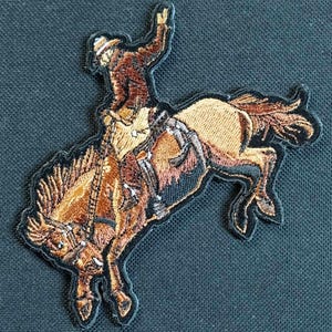May include: An embroidered patch depicting a cowboy riding a bucking horse. The cowboy is wearing a brown hat and jacket, while the horse is brown with a white mane and tail.