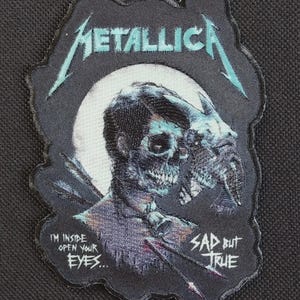 Può includere: Una toppa ricamata nera con il logo Metallica in azzurro. Il design raffigura una figura simile a un teschio con una campana al collo, frecce e una luna bianca sullo sfondo. Testo: I'M INSIDE OPEN YOUR EYES... SAD BUT TRUE.