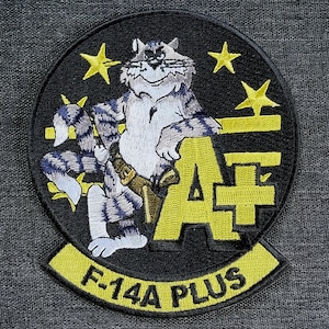 May include: Embroidered patch featuring a cartoon cat in a military-style design. The cat is gray and white with blue stripes, wearing a belt and holding a tool. The patch has yellow stars, the letters "A+", and the text "F-14A PLUS" on a black background.