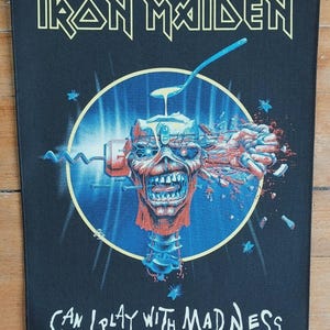 May include: Black rectangular patch featuring the text "IRON MAIDEN" in yellow, above a circular illustration of a skull with a spoon and electrical components. The text "CAN I PLAY WITH MADNESS" is below the illustration.