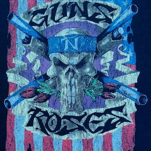 May include: Black t-shirt featuring a distressed graphic of a skull with crossed guns, roses, and the text "GUNS N' ROSES". The design incorporates elements of the American flag with red, white, and blue colors. The shirt has a vintage, worn look.