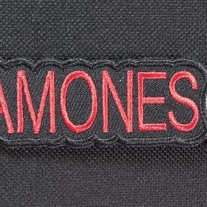 May include: A black patch with the word "RAMONES" embroidered in red, outlined in black. To the right is a circular patch with a white design and the words "JOHNNY", "TOMMY", "DEE DEE", and "JOEY" around the edge.