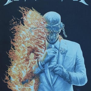 May include: A black t-shirt featuring the word MEGADETH in white lettering at the top. The graphic shows a skull-faced figure in a light blue suit and tie, wearing futuristic goggles and a chain, with bright orange flames engulfing the right side.