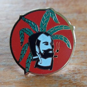 May include: A red and green enamel pin featuring a cartoon man with a beard and a pipe. He is wearing a green leafy crown and smoking a pipe.
