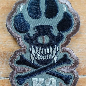 May include: A green and brown embroidered patch with a black paw print, skull, and crossbones. The text "K9" is embroidered in white.