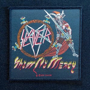 Könnte beinhalten: Ein schwarzer quadratischer Aufnäher mit dem Wort "SLAYER" in Weiß, Rot und Orange in einem Pentagramm. Rechts ist eine Illustration eines Kriegers mit einem Schwert. Der Text "SHOW NO MERCY" steht darunter, mit "©2001 SLAYER" unten.