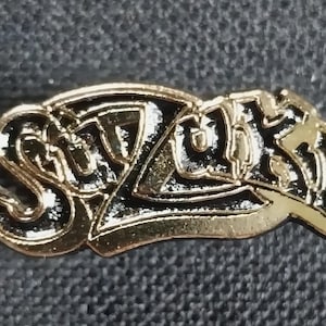 May include: A gold-colored metal pin with the word "SUZUKI" in black lettering. The letters are stylized and have a 3D effect.
