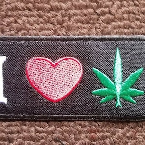May include: Black embroidered patch with the text "I love" followed by a red heart and a green marijuana leaf.