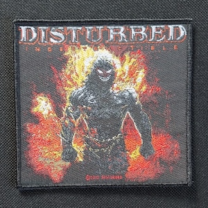 May include: A square patch with the band name "DISTURBED" at the top. Below is an illustration of a dark figure engulfed in flames. The figure has red eyes and is surrounded by orange and yellow flames. The word "Indestructible" is also visible.