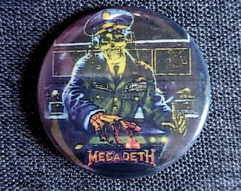 May include: Round button featuring a skeletal figure in a military uniform, wearing a hat and headset, operating a control panel. The button has the band name "MEGADETH" in a stylized font at the bottom. The color scheme is dark with blue, green, and yellow accents.