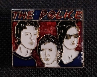 May include: A gold-colored pin with a red, white, and blue image of the band The Police. The band members are depicted in a cartoon style with black hair and white faces. The band's name is written in red and white letters on a blue background.