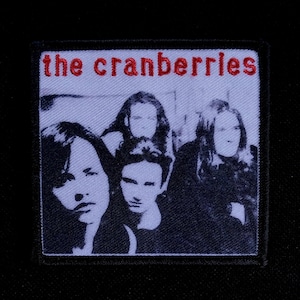 May include: A square patch featuring a black and white image of four people. The text "the cranberries" is embroidered in red at the top. The patch is likely for a jacket or bag, showcasing a band.