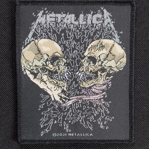 May include: Black rectangular patch featuring the band name "METALLICA" in a distressed, stylized font above two detailed skull illustrations. The skulls are light beige with dark shading. The bottom of the patch reads "©2021 METALLICA".