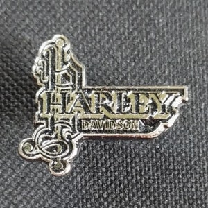 Può includere: Spilla Harley-Davidson color argento con la scritta "HARLEY DAVIDSON" e il numero "15" in un design stilizzato. La spilla, di forma rettangolare, presenta decorazioni e si staglia su uno sfondo scuro.