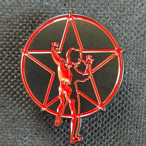 May include: A red and black metal pin featuring a five-pointed star with a figure inside. The figure is in a pose with arms raised.