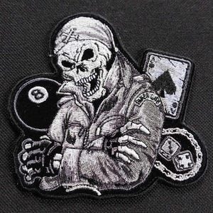Pode incluir: Um remendo bordado em preto e branco de uma caveira com uma bandana na cabeça e segurando uma bola de bilhar. A caveira está vestindo uma jaqueta com o texto "Poker Club" na manga. O remendo também inclui um cartão de jogo e uma corrente com dados.