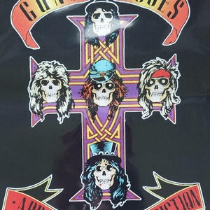 以下が含まれることがあります： 十字架と5つのドクロの顔を持つ、白黒のガンズ・アンド・ローゼズのロゴ。上部に黄色のバナーに「ガンズ・アンド・ローゼズ」、下部に赤いバナーに「アペタイト・フォー・デストラクション」の文字が書かれている。