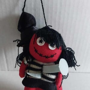 May include: A handmade red doll with black hair and a stitched smile. The doll wears a black and white striped shirt and a black skirt. It holds a small, gray knife and is suspended by a black string.