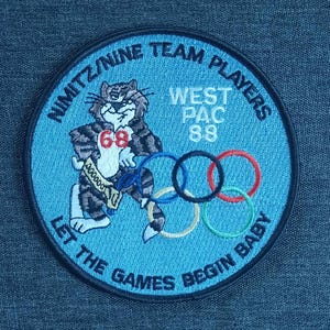 May include: A blue and black embroidered patch with a cartoon cat wearing a shirt with the number 68. The text "Nimitz Nine Team Players West Pac 88 Let the Games Begin Baby" is around the edge of the patch.