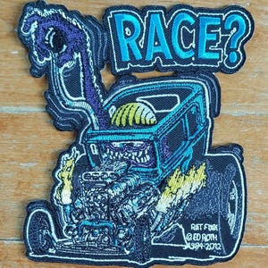 May include: Embroidered patch depicting a cartoon hot rod with a monster engine and the text "RACE?". The patch has a black background with blue, yellow, and purple accents. The text "RAT FINK @ ED ROTH 1994-2012" is also included.