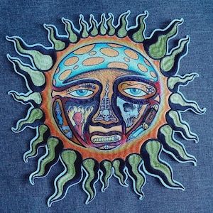May include: Embroidered patch depicting a stylized sun with a face. The sun has a blue and orange face with detailed imagery, including a skull. The sun's rays are green and yellow. The patch is likely for clothing or accessories.