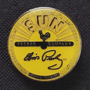 May include: A round, yellow pin featuring the text "SUN RECORD COMPANY" with a rooster graphic. The pin also includes Elvis Presley's signature and the text "MEMPHIS, TENNESSEE". The design is set against a musical note background.