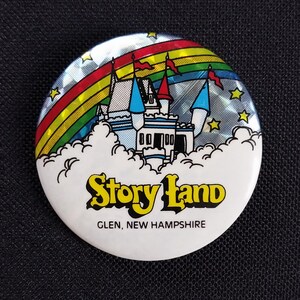 May include: A round, white Story Land button with a holographic background. The button features a colorful rainbow, a white castle with blue and red accents, and the text "Story Land, Glen, New Hampshire."