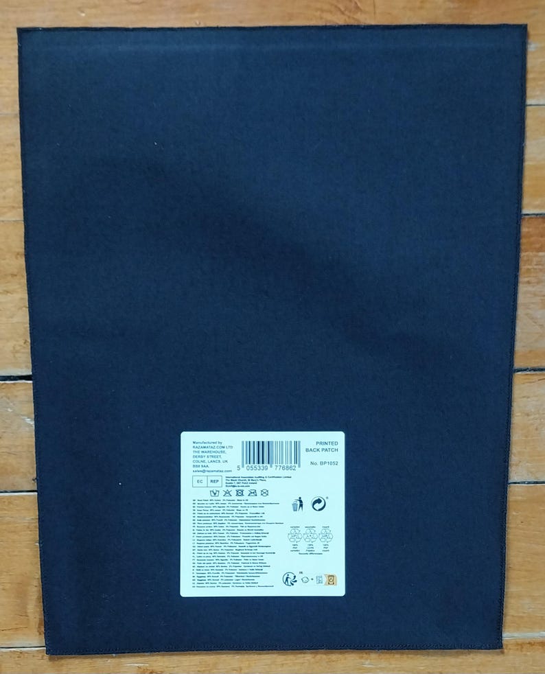 May include: A dark navy blue rectangular patch with a white label in the upper right corner. The label contains text, a barcode, and various symbols. The patch is lying on a wooden surface.