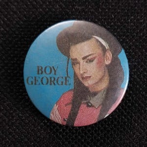 May include: A blue and white button featuring a picture of Boy George, a singer, wearing a black hat and a pink shirt. The text "Boy George" is printed on the button.