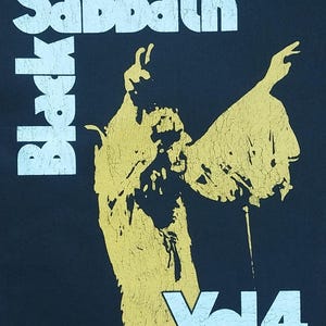 Pode incluir: Camiseta preta com um gráfico Black Sabbath Vol 4. O design apresenta o nome da banda em branco, empilhado verticalmente, com uma figura amarela e o texto "Vol 4" em branco. Uma peça de vestuário com tema musical.