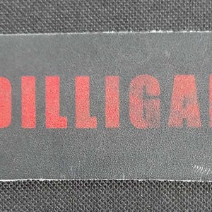 May include: A rectangular patch with rounded corners, featuring the red text "DILLIGAF" on a dark gray background. The patch has a white border and is likely made of fabric or a similar material.