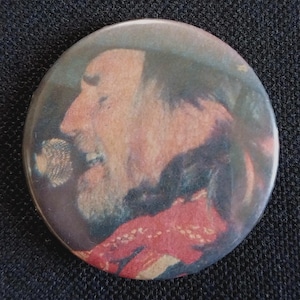 May include: Round button pin featuring a profile portrait of a man with a beard singing into a microphone. The image has a vintage, slightly faded appearance, with a red and blue color scheme. The pin is likely a collectible or a piece of memorabilia.