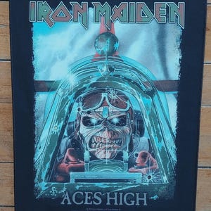 Puede incluir: Parche negro con el logotipo de Iron Maiden y la ilustración del sencillo "Aces High". La imagen muestra a Eddie, la mascota de la banda, en la cabina de un avión de combate. El texto "ACES HIGH" se muestra en la parte inferior.