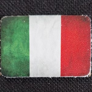 May include: A rectangular sticker featuring the Italian flag. The flag is divided into three vertical stripes: green on the left, white in the center, and red on the right. The sticker has rounded corners and a slightly distressed appearance.