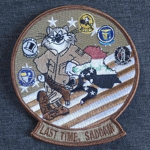 May include: A brown and tan embroidered patch featuring a cartoon cat in a military uniform holding a gun. The cat is standing on a striped background with the text "Last Time Saddam" at the bottom.